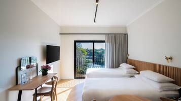 A modern hotel room with a large bed, a desk with a flat-screen TV, and a balcony with a view of greenery.