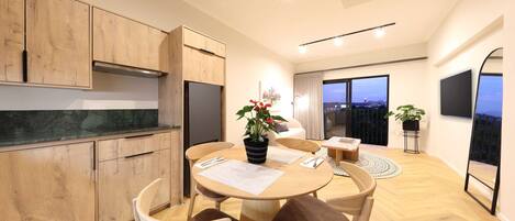 A modern kitchen with wooden cabinets, a dining area with a round table and chairs, and a balcony with a view.