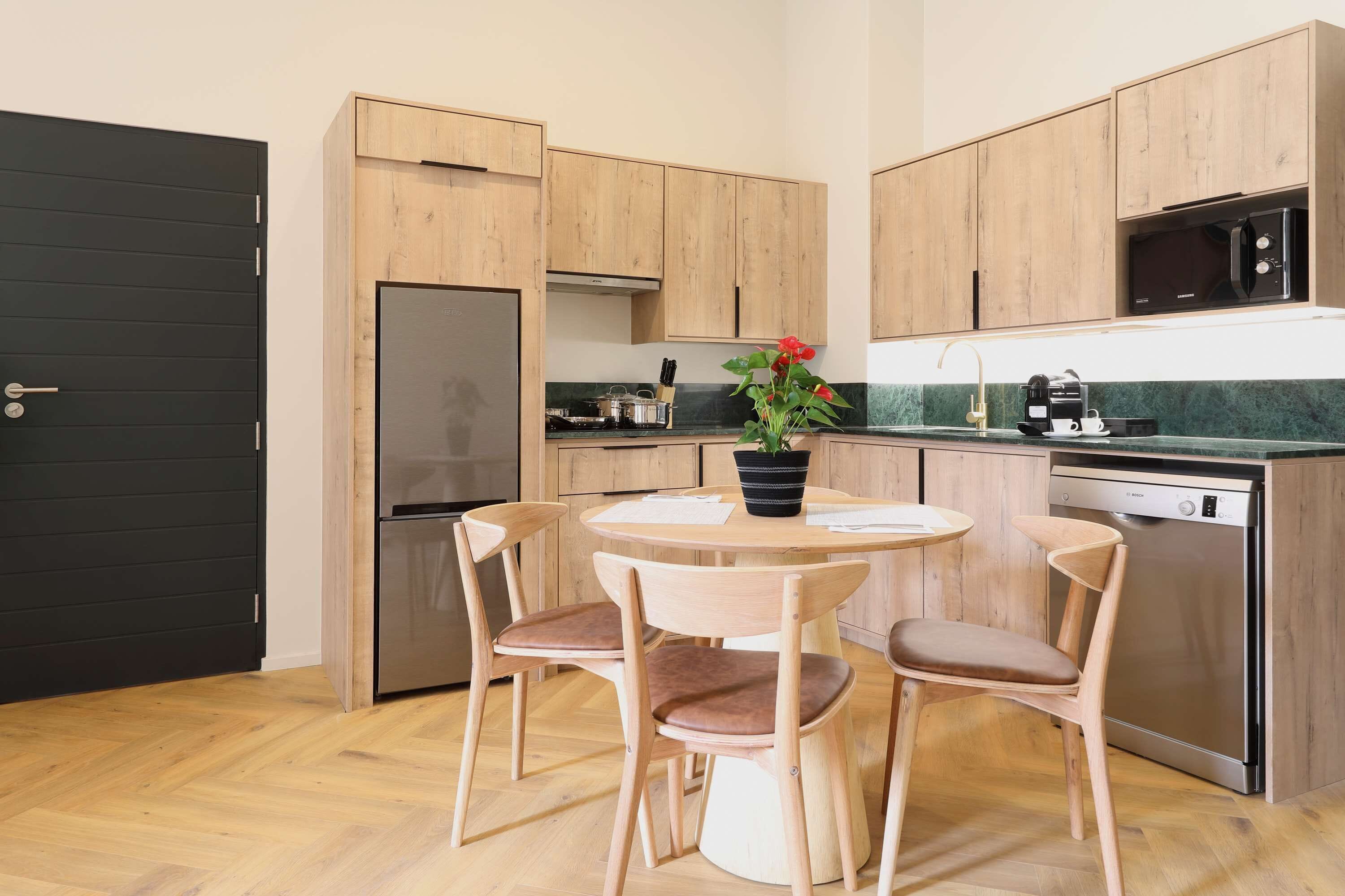 Cocina compacta con armarios de madera, un refrigerador negro, microondas y cafetera. Hay una mesa redonda con dos sillas y una planta en maceta.