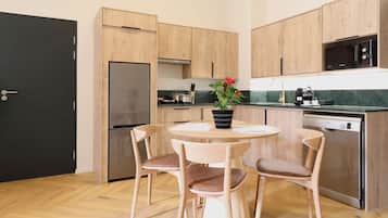 Cozinha compacta com armários de madeira, uma geladeira preta, micro-ondas e cafeteira. Há uma mesa redonda com duas cadeiras e um vaso com planta.