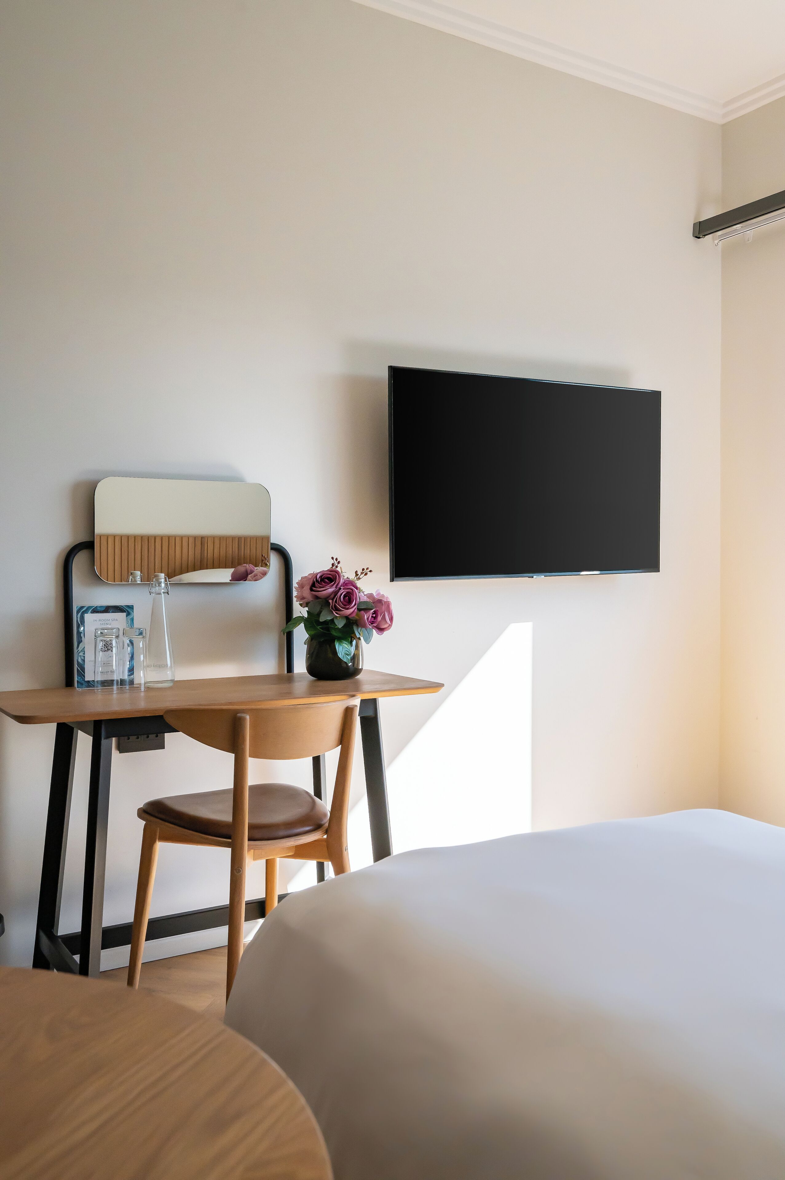 A hotel room with a bed, a desk with a chair, a mirror, a flat-screen TV, and a vase with flowers.