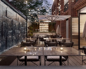 An outdoor dining area with tables and chairs, a bar with bottles and glasses, and a brick wall.