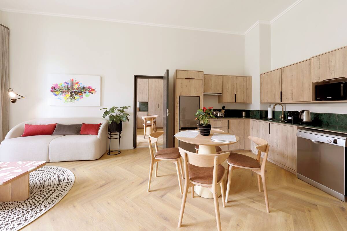 Una cocina moderna con armarios de madera, horno empotrado y lavavajillas. Hay una zona de comedor con una mesa redonda y sillas.