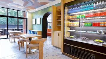 Un café con mesas y sillas de madera, un refrigerador lleno de bebidas y vistas amplias a través de grandes ventanales.