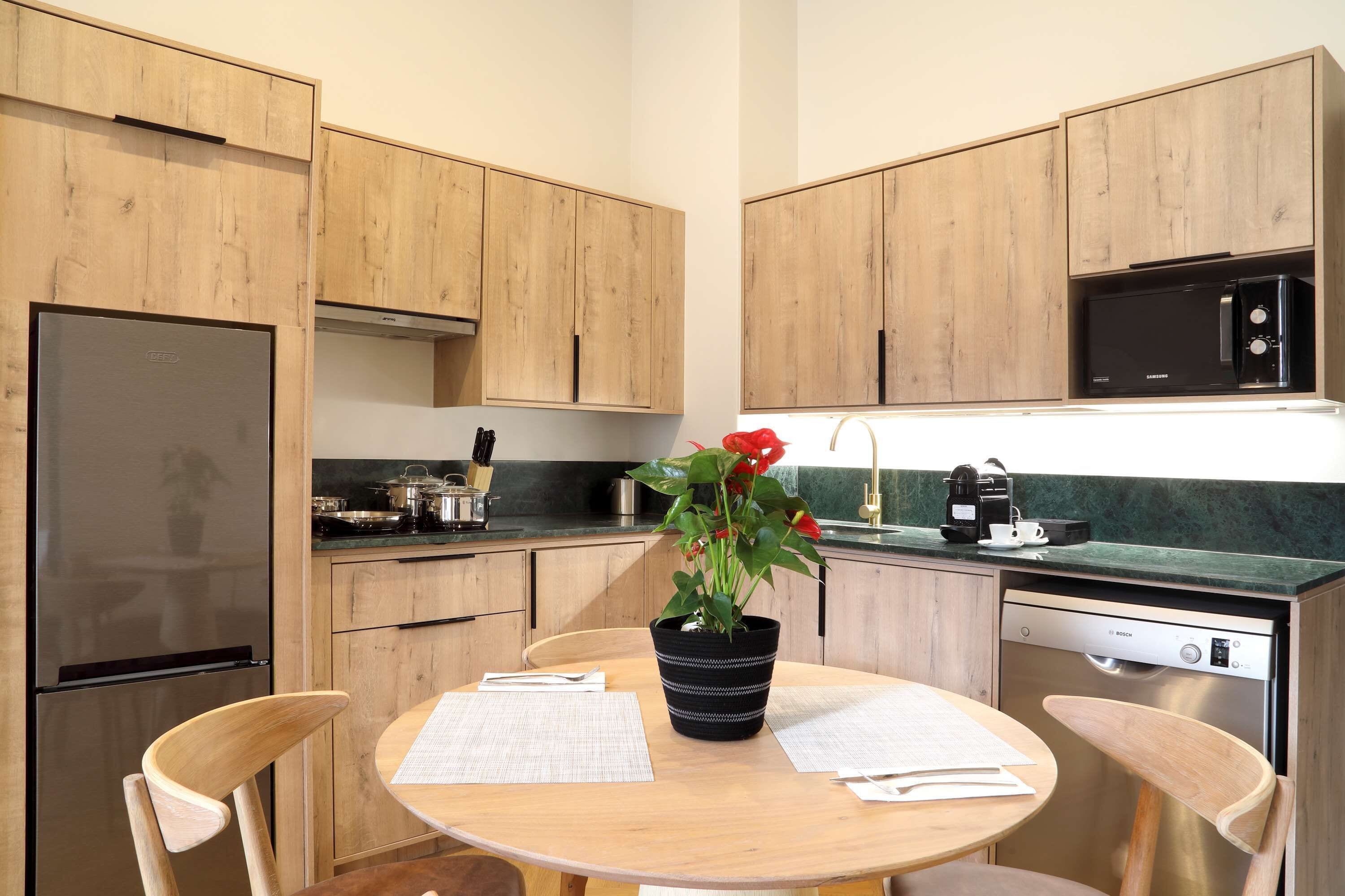Une cuisine moderne avec des meubles en bois, un réfrigérateur en acier inoxydable, un lave-vaisselle Bosch et une table sur laquelle se trouvent des papiers et un pot de fleurs.
