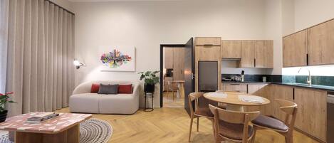 A modern living room with a sofa, coffee table, and dining area. A kitchen with wooden cabinets and appliances is visible in the background.