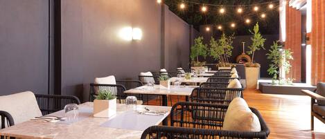 Un área de comedor al aire libre con varias mesas y sillas, luces colgantes y plantas en macetas.