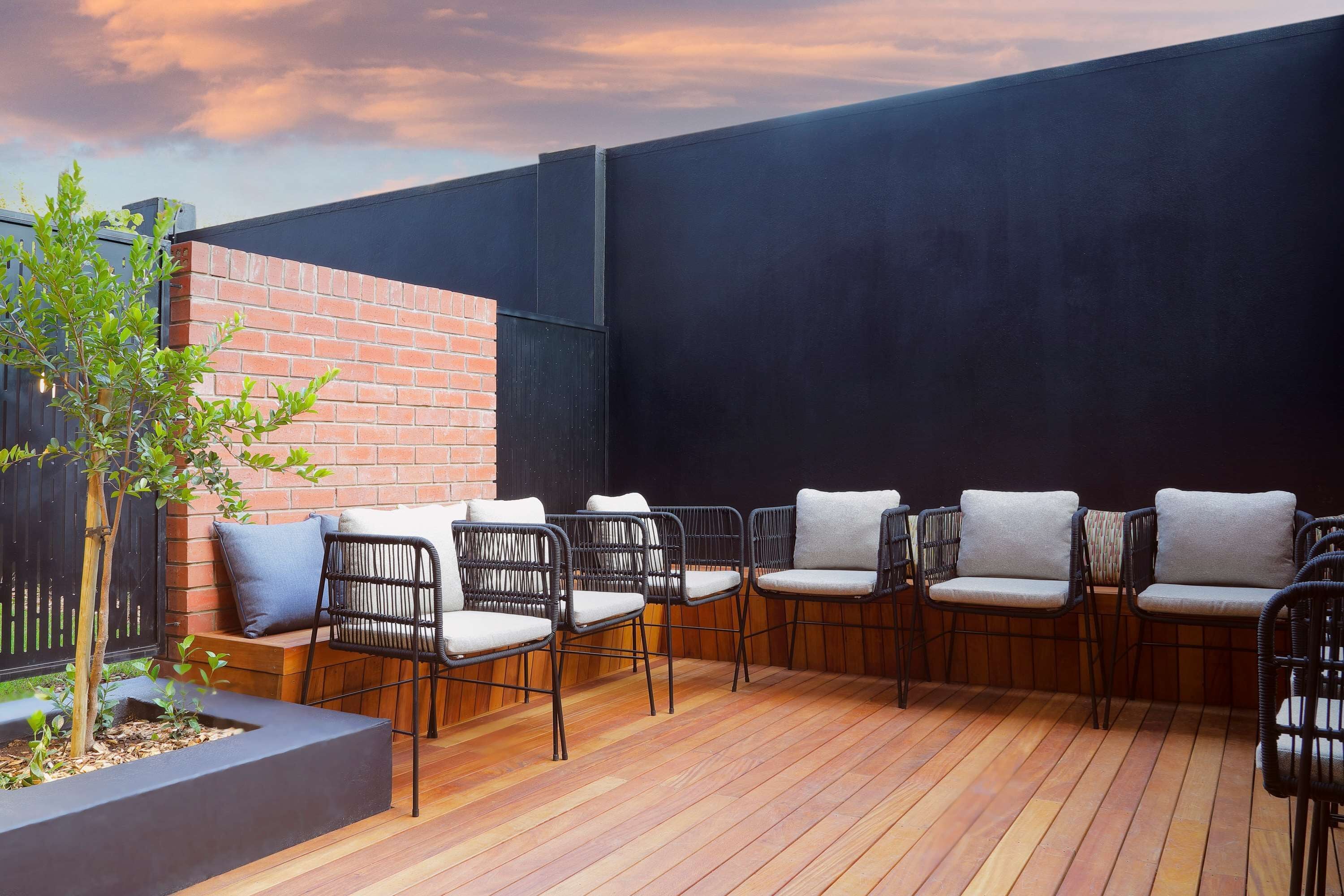 Un patio con suelo de madera, sillas de metal negro y un muro de ladrillo.