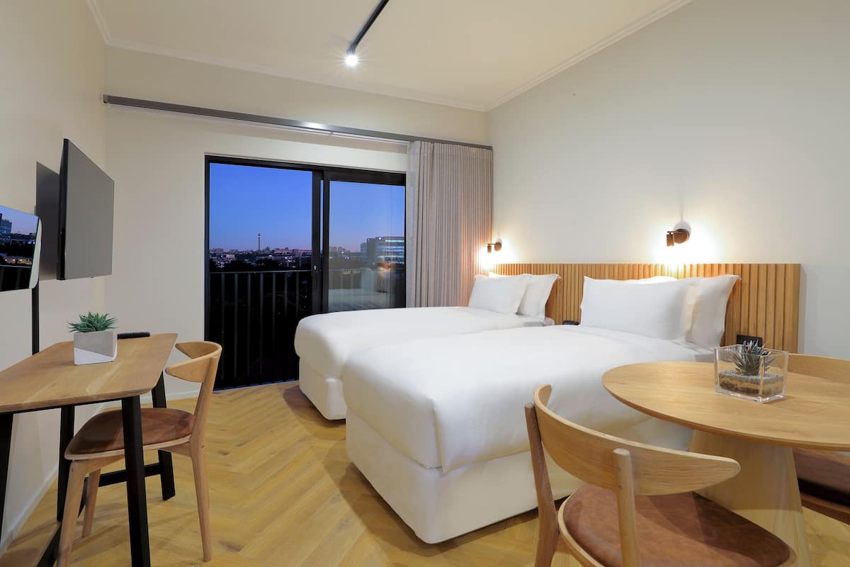 Habitación de hotel con una cama grande, un escritorio con silla, una mesita con un vaso, y vista a la ciudad al anochecer.