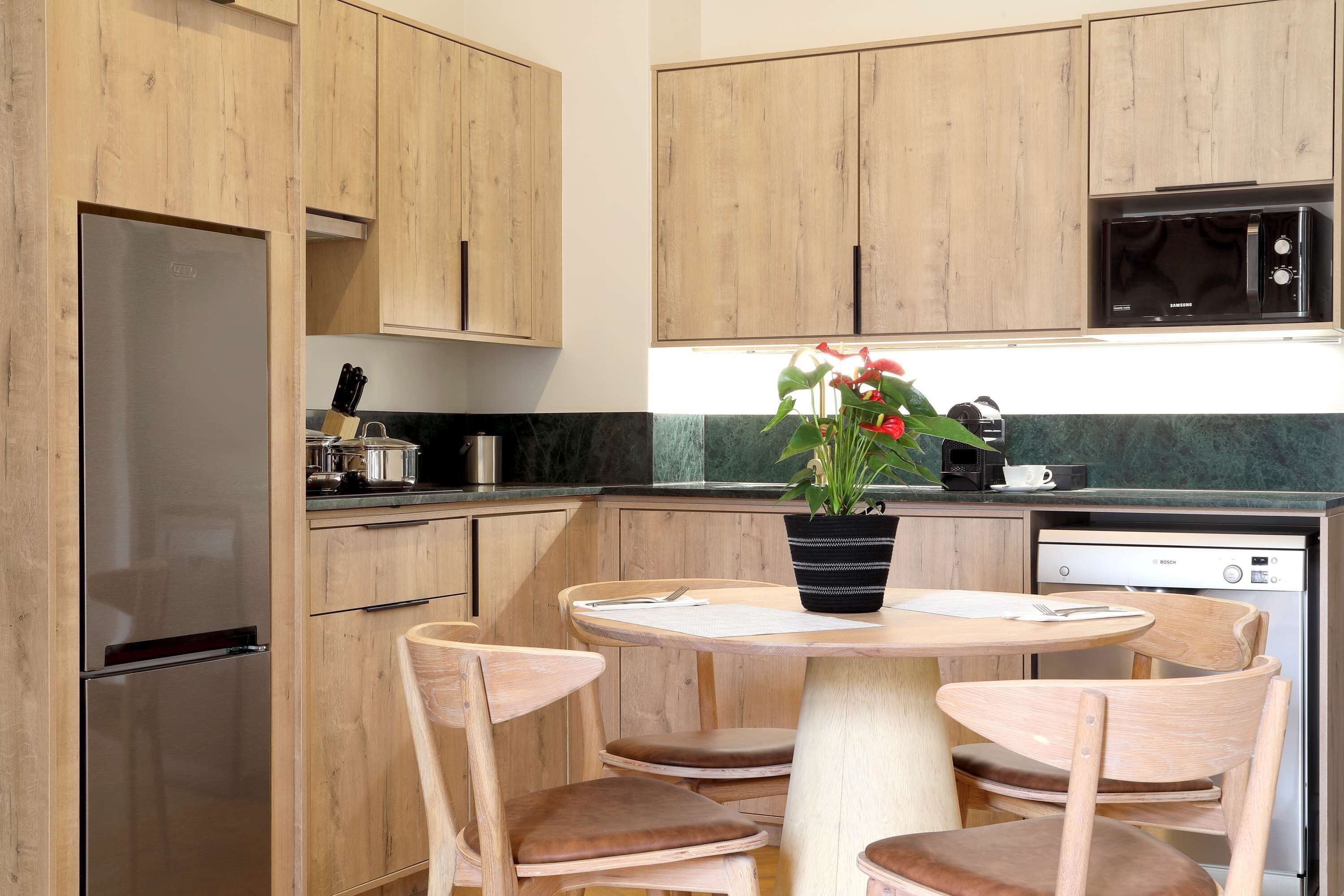 A modern kitchen with wooden cabinets, a stainless steel refrigerator, a microwave, and a dishwasher. There is a round table with chairs and a potted plant on the counter.