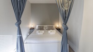 Ein ordentlich bezogenes Bett mit Kissen und einem Nachttisch mit Lampe, in einem Zimmer mit hellen Wänden und an den Ecken gebundenen Vorhängen.