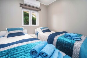 Ein Zimmer mit zwei Betten, jeweils mit blau-weißer Bettwäsche, einem Fenster mit Blick ins Grüne und einer Klimaanlage über dem Fenster.
