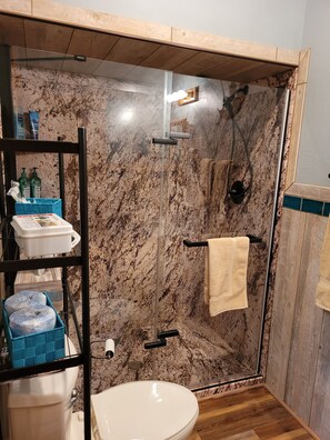 Un baño con ducha envuelta en vidrio, inodoro, lavabo y estante con artículos de tocador.