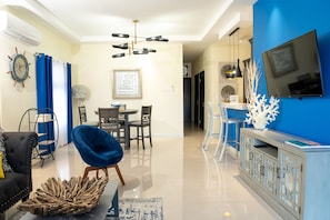 Un salon moderne comprenant un coin repas, un fauteuil bleu, une table basse en bois et un téléviseur fixé à un mur bleu.