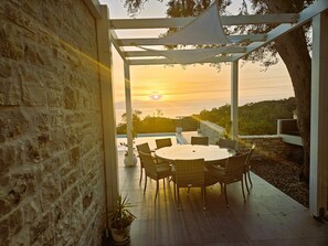 A covered outdoor seating area with a table and chairs, overlooking a pool and a sunset.