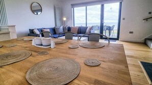 A modern living room with a wooden dining table, blue sofas, and a large window offering a view of the sea.