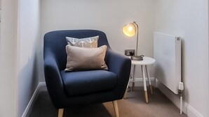 A small, cozy room with a sloped ceiling and a skylight, featuring a dark blue armchair with a cushion, a small table with a lamp, and a radiator.