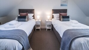 A modern, well-lit room with two single beds, each with a nightstand and a lamp. The room has a sloped ceiling and a framed picture on the wall.