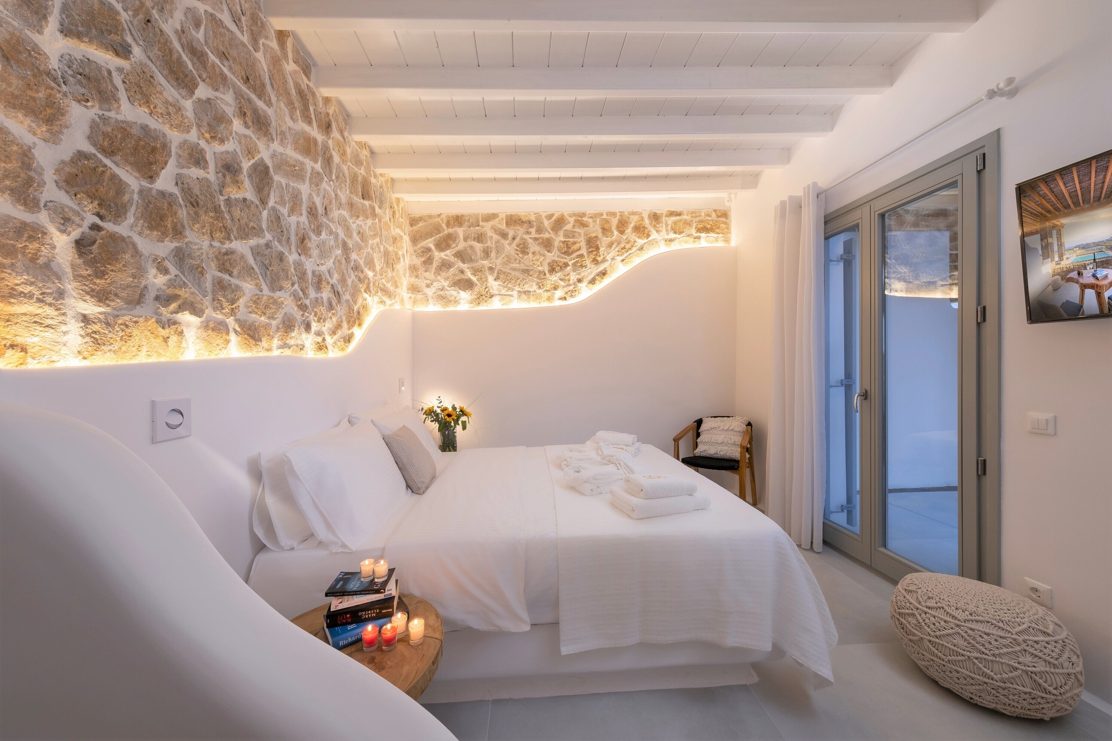 Une chambre moderne avec un mur en pierre, un lit recouvert de linge de maison blanc, une petite table de chevet avec des livres et des bougies, et une vue à travers une porte-fenêtre coulissante.