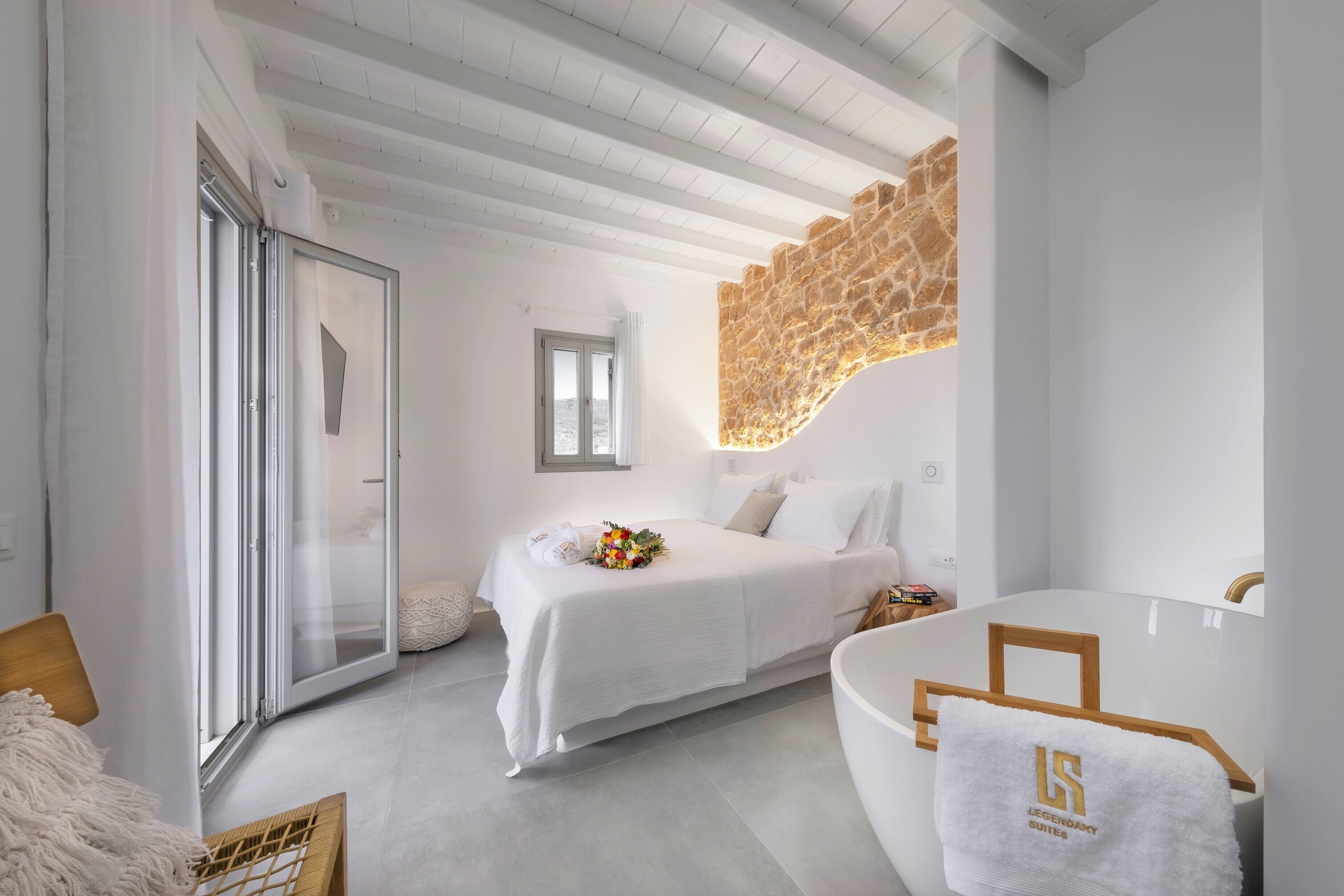 A modern bedroom with a large bed, a glass door, a stone accent wall, and a bathtub with a towel.