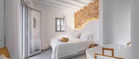 A modern bedroom with a large bed, a glass door, a stone accent wall, and a bathtub with a towel.