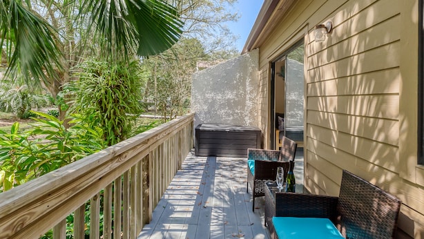 A wooden deck with outdoor furniture, a glass door leading to a room, and surrounding greenery.
