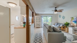 A modern living room with a grey sofa, a wooden coffee table, and a TV. There is a kitchen area with a fridge and a counter.