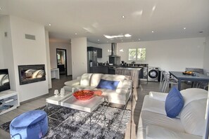 A modern living room with a sofa, coffee table, and a TV. There is a kitchen area with a stove, sink, and dining table.
