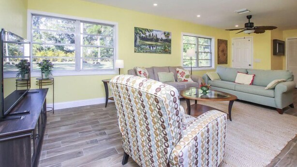 A living room with a sofa, armchair, coffee table, and a TV on a stand. There are windows, a ceiling fan, and a door. - Pine Island Tropical Retreat 1 (Saint James City)