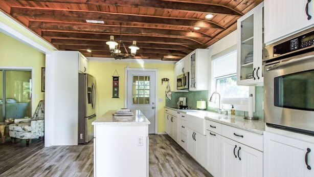 Una cocina con armarios blancos, una isla central, techo de madera y una puerta que da a otra habitación.