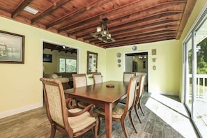 A dining area with a wooden table and chairs, a ceiling fan, and a view through large windows.