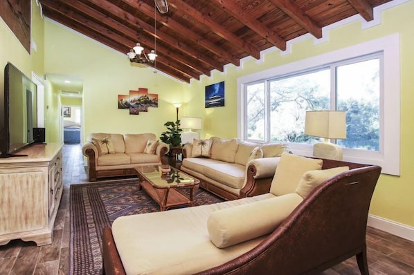 A living room with a wooden ceiling, a beige sofa, a coffee table, and a large window with a view of trees.