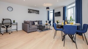 A modern living room with a grey sofa, a dining table with blue chairs, a black office chair, and a small side table with a plant. - Watford Cassio Deluxe - Modernview Serviced Accommodation (Watford)