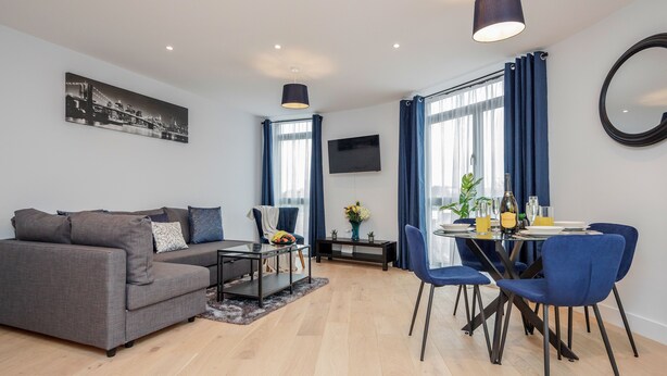 A modern living room with a grey sofa, a dining table with blue chairs, a flat-screen TV, and a large window with curtains. - Watford Cassio Deluxe - Modernview Serviced Accommodation (Watford)