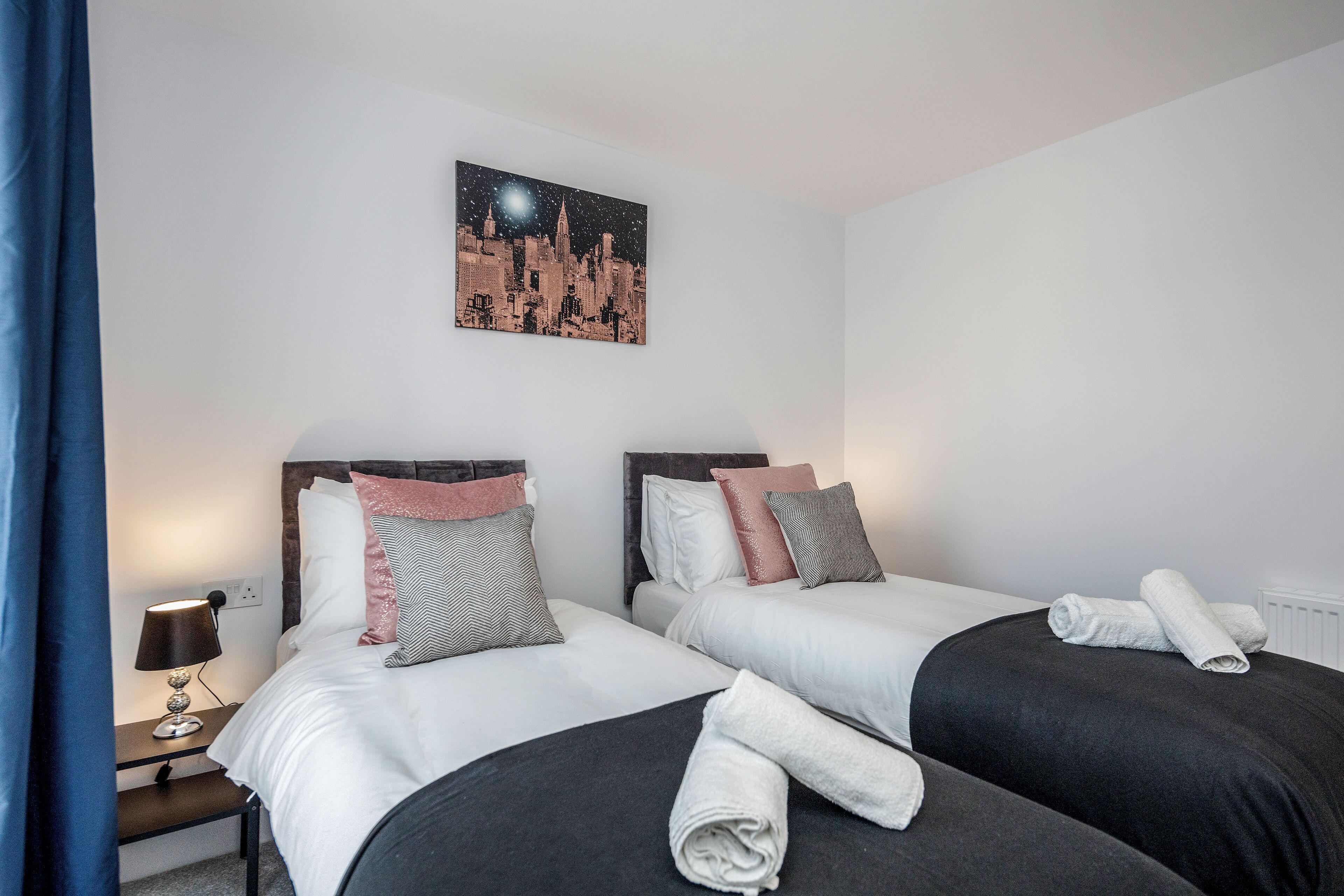 Ein Hotelzimmer mit zwei Betten, einem Nachttisch mit Lampe und einem Stadtbild an der Wand.