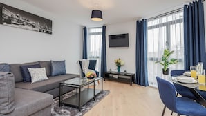 A modern living room with a grey sofa, a glass coffee table, and a dining area with blue chairs and a small table. - Watford Cassio Deluxe - Modernview Serviced Accommodation (Watford)