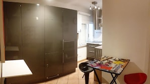 A room with a large wardrobe, a small table with a magazine, a red chair, and a kitchenette in the background.