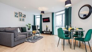 A modern living room with a grey sofa, a dining table with green chairs, a TV mounted on the wall, and a large window with curtains. - Watford Cassio Luxury - Modernview Serviced Accommodation (Watford)
