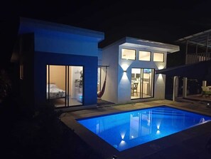 A modern house with a swimming pool at night, featuring large windows and a hammock.