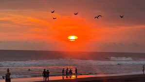 Een zonsondergang boven de zee met mensen op het strand en vogels die vliegen.