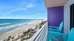 A balcony with turquoise chairs overlooking a beach with umbrellas and palm trees.