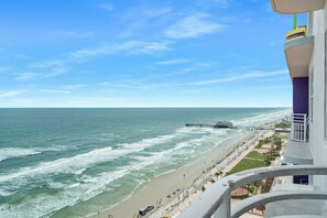 Een kustgezicht met een steiger die zich uitstrekt over de oceaan, een strand met mensen en parasols, en een gebouw met een balkon met uitzicht op het tafereel.