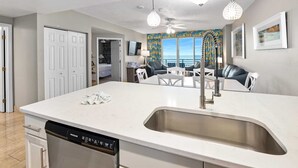 A modern kitchen with a central island, stainless steel sink, and a dishwasher. There is a dining area with a table and chairs, and a view of the ocean through large windows.