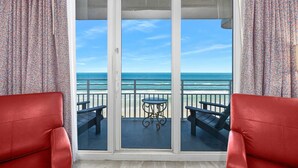 A room with a view of the ocean through a sliding glass door, featuring two red chairs and a small table.