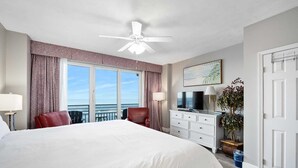 A hotel room with a large bed, a ceiling fan, a TV, a dresser, and a view of the ocean.