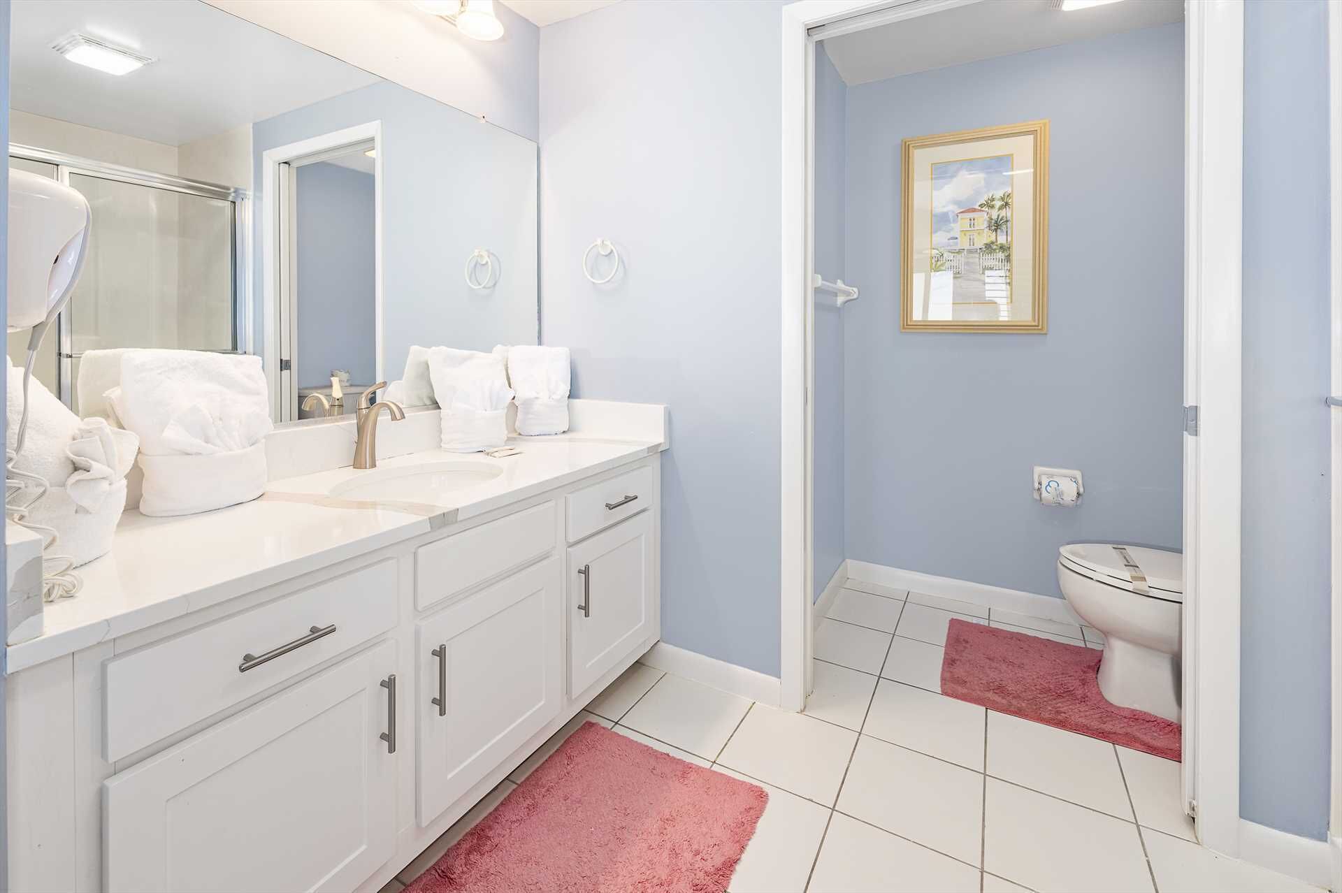 A bathroom with a white vanity, a large mirror, a sink, and a toilet. There is a framed picture on the wall and a pink rug on the floor.
