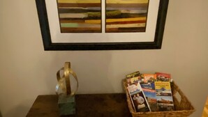 Ein gerahmtes abstraktes Gemälde an einer Wand, ein kleiner Tisch mit einer Buchkorb und eine dekorative Vase.