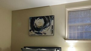 Ein Schlafzimmer mit einem Bett, einem Nachttisch mit Lampe, einem an der Wand hängenden abstrakten Gemälde und einem Fenster mit Jalousien.