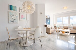 A modern living room with a round dining table, white chairs, a sofa, and a coffee table. There are framed artworks on the walls and a large window providing natural light. - Jardines de Calahonda III | Welcoming Apartment with Sea Views & Terrace (Mijas Costa)