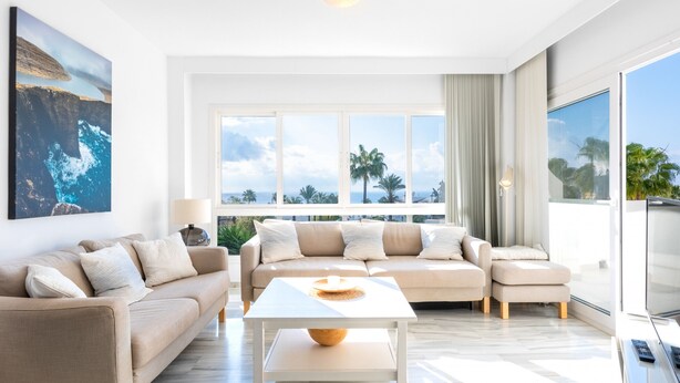 Un salon moderne avec un ensemble de canapés beige, une table basse blanche et de grandes fenêtres donnant sur des palmiers et l’océan.